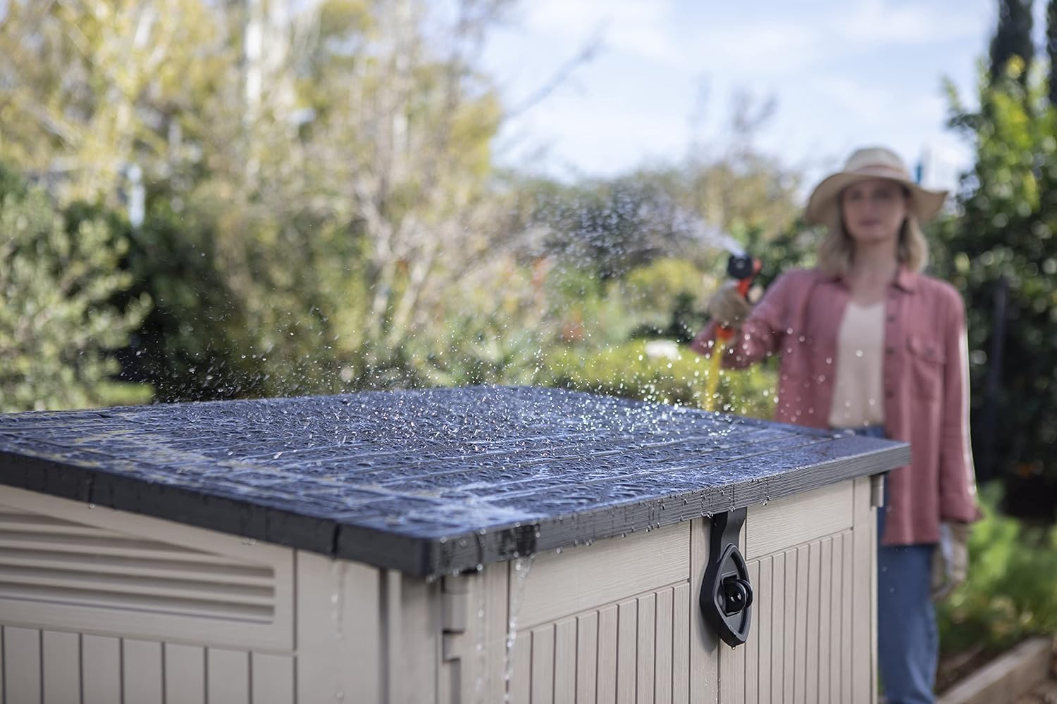 Cobertizo de jardín exterior Store It out Midi 880 litros, topo/beige. Vista en exterior con mujer mojando con una manguera. Keter | maxjardin